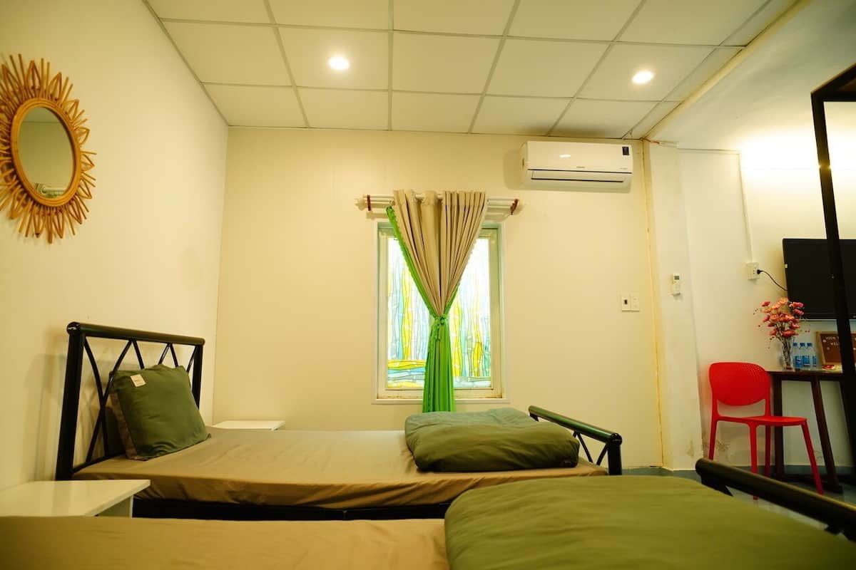 Una habitación con dos camas individuales, una mesita, una silla, un ventanal con cortinas y un equipo de aire acondicionado.