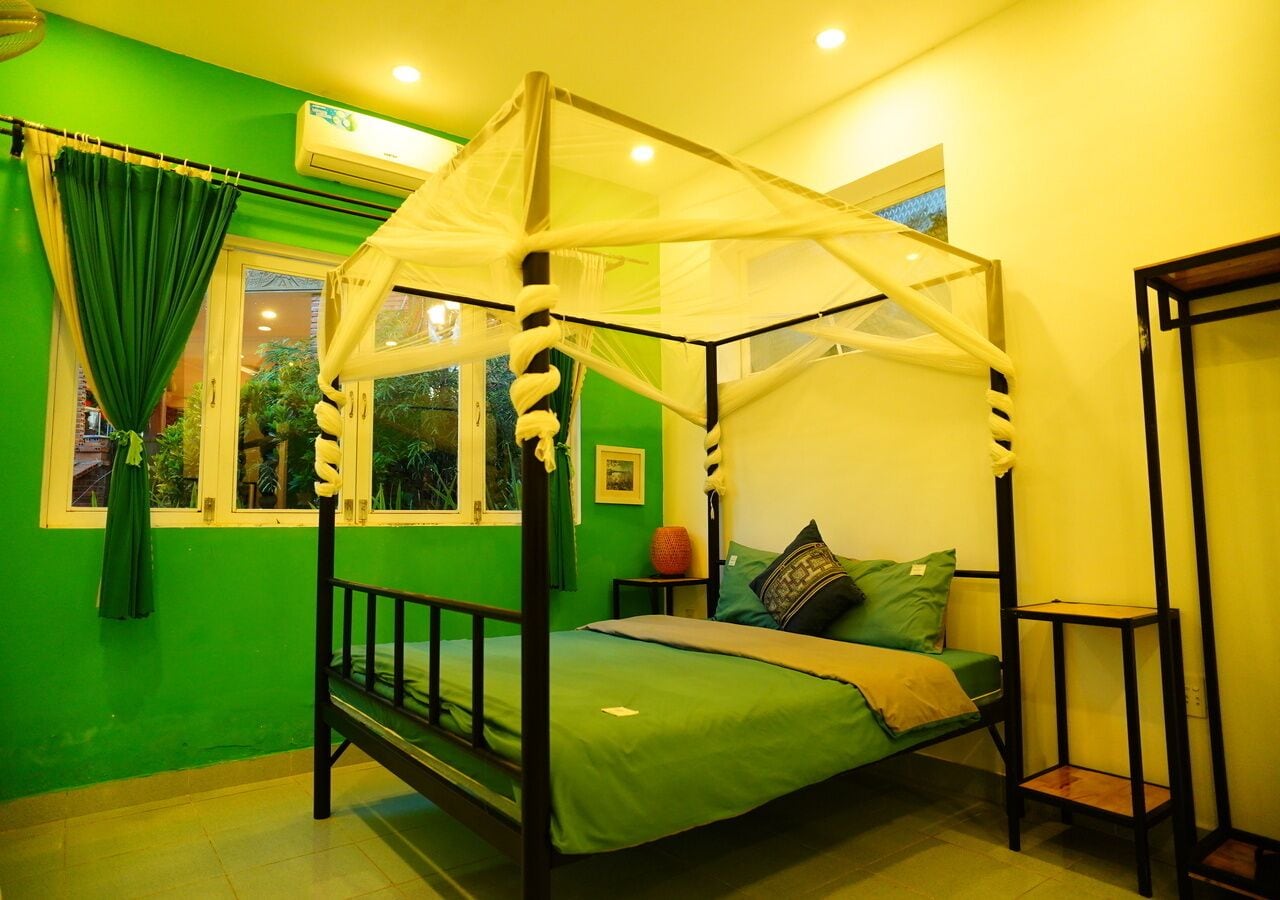 A room with a four-poster bed, green walls, a window with a view of greenery, and a small table with a vase.