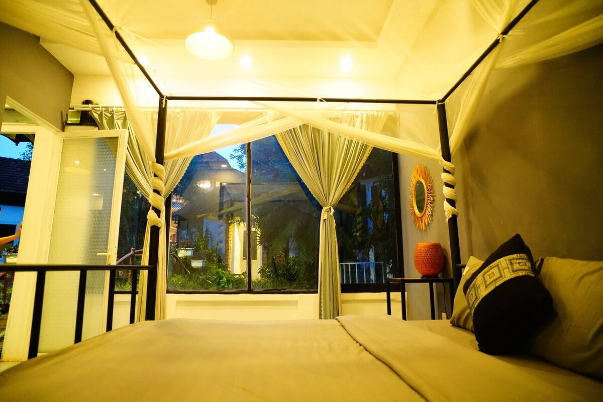 Una habitación con cama, una ventana con cortinas, una mesita con un florero y un adorno en la pared.