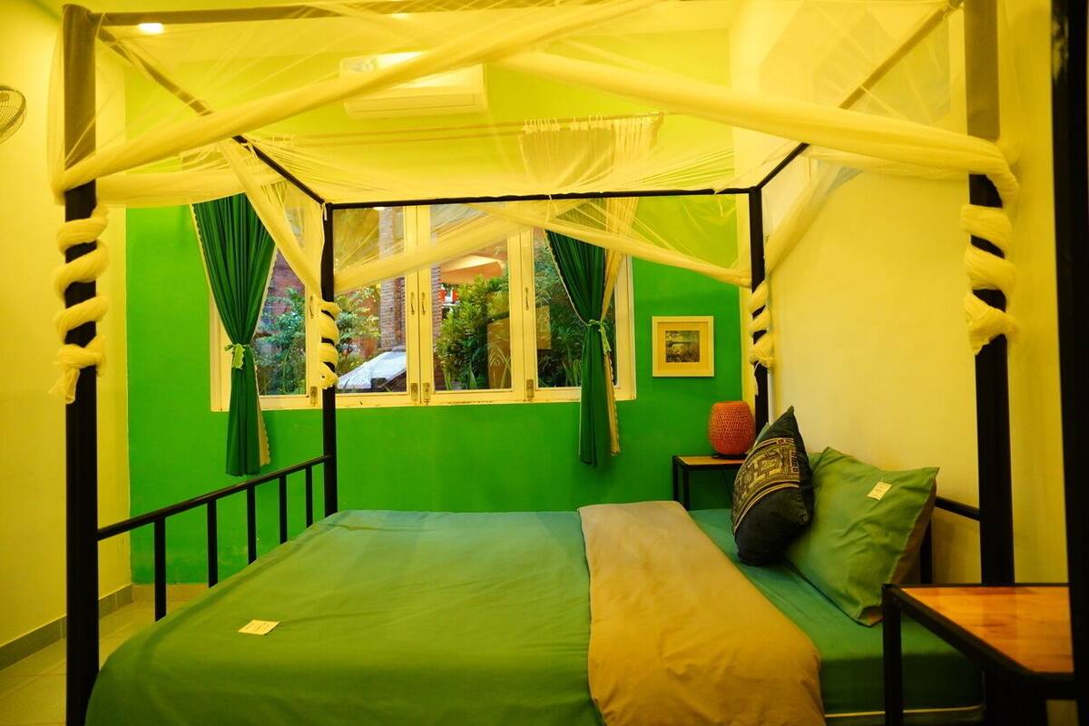 Habitación con cama bajo un dosel, paredes verdes, una ventana con cortinas y una pequeña mesa con un cojín.