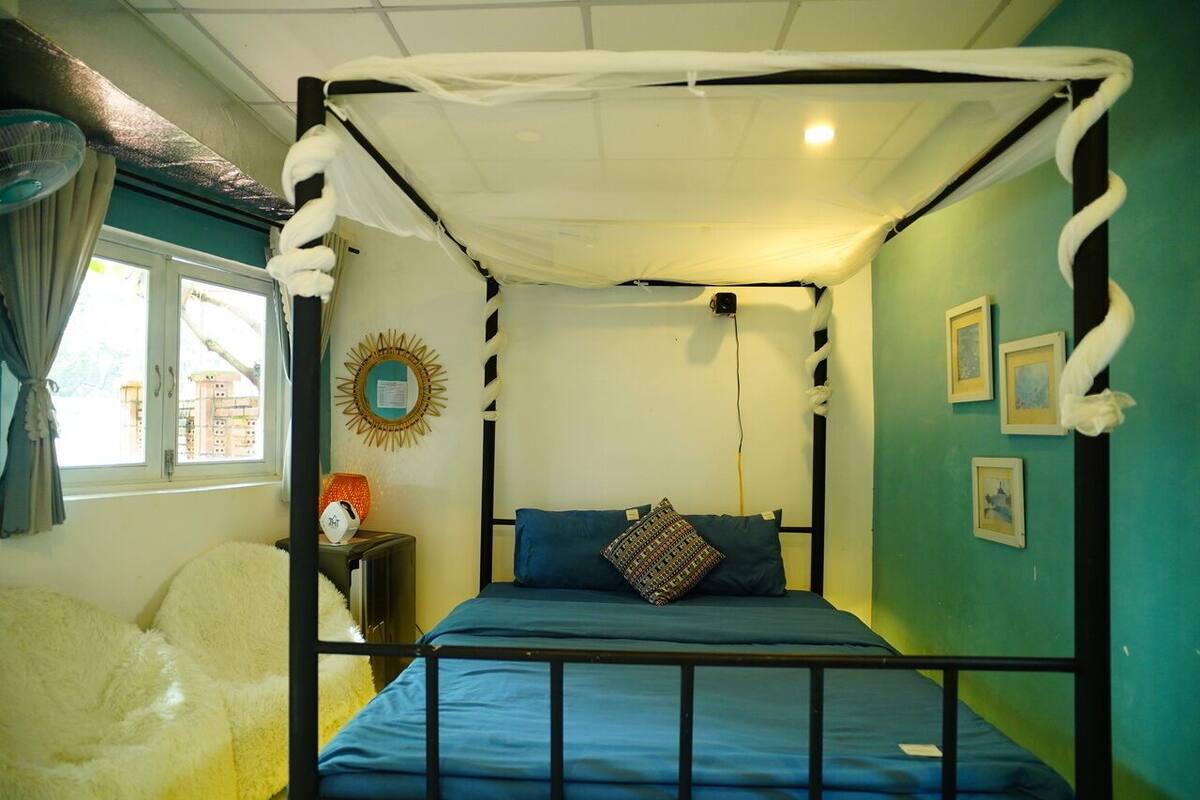 Una habitación con una cama con dosel, una mesita de noche pequeña con una lámpara y una ventana con cortinas.