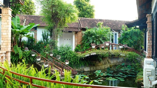 A traditional building with a brick exterior, a small bridge over a pond with lily pads, and lush greenery surrounding the area. - Vuon Thu Do Homestay (Buon Ma Thuot)