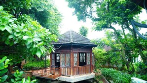 A wooden structure with a tiled roof, surrounded by lush greenery and a water body. - Vuon Thu Do Homestay (Buon Ma Thuot)