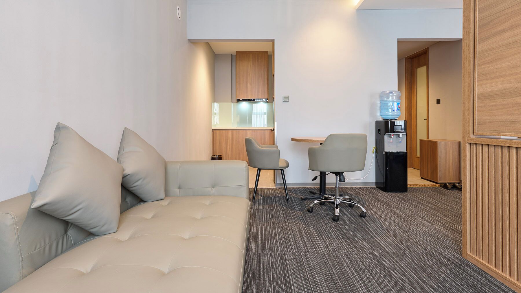 A hotel room with a sofa, two chairs, a small table, a water dispenser, and a kitchenette.