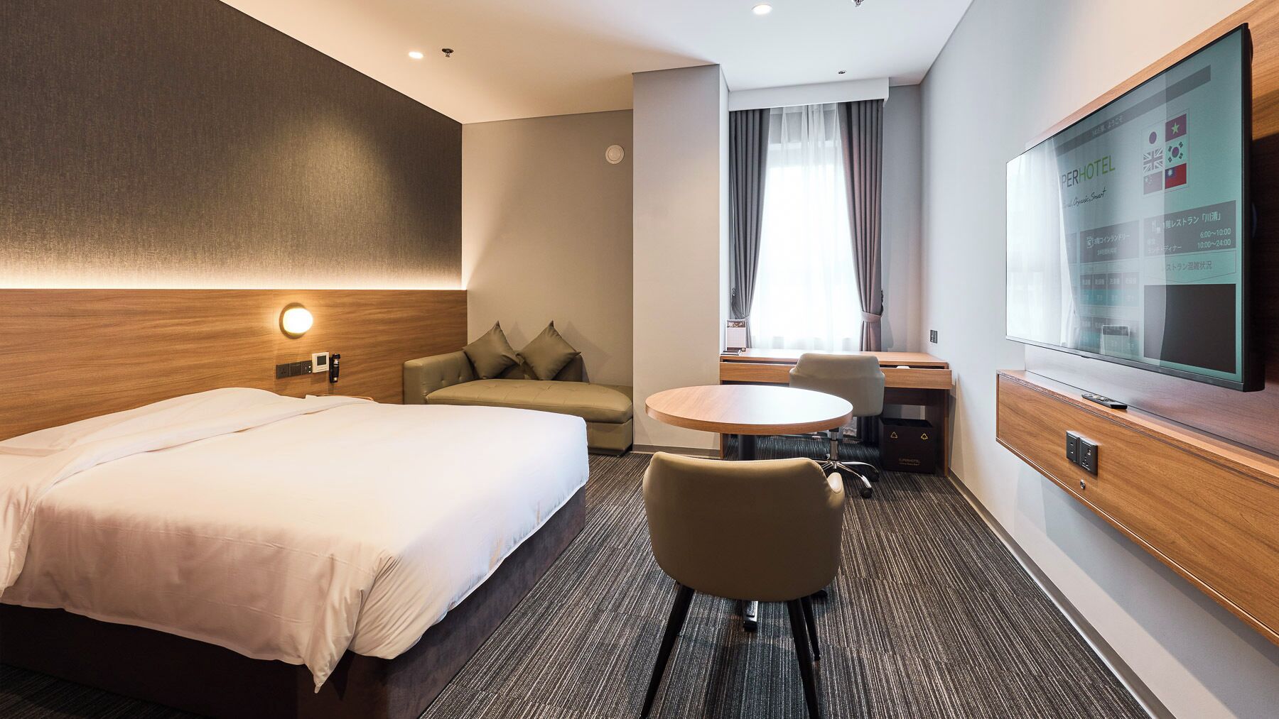 Habitación de hotel con cama, sofá, escritorio con silla, televisor y una pantalla mural que muestra información de la habitación.