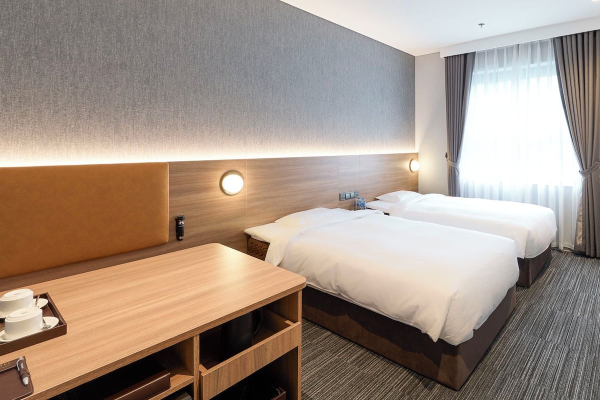 Habitación de hotel con dos camas, un escritorio con cafetera, un televisor y un ventanal con cortinas.