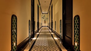Un long couloir étroit, doté d’un sol à motifs et bordé de portes ornées de panneaux de verre décoratifs, mène à une pièce où se trouve une plante.