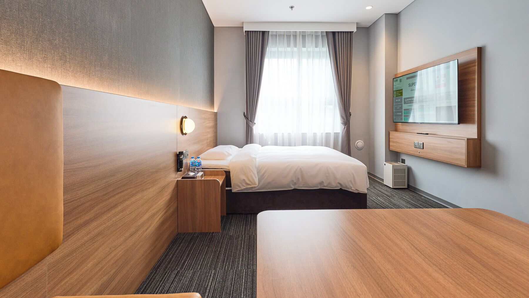 Una habitación de hotel con cama, escritorio con lámpara, televisor en la pared y ventana con cortinas.