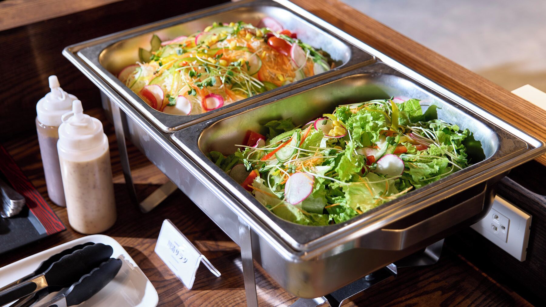 A buffet with two metal serving trays containing salads, a bottle of dressing, and a pair of scissors.
