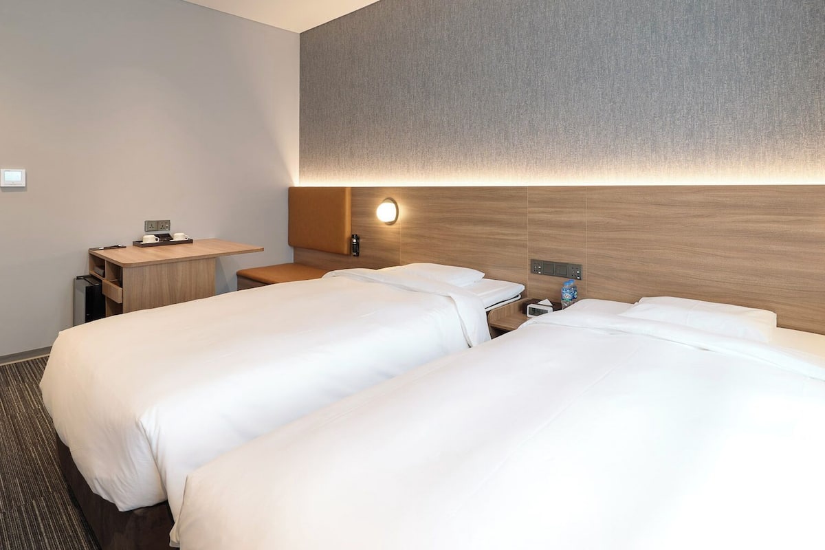 Habitación de hotel con dos camas, un escritorio, una silla, una mesita pequeña y una lámpara fijada a la pared.
