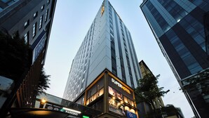 Un immeuble de grande hauteur doté d’une façade moderne en verre, un hôtel avec une enseigne lumineuse, et un pont piétonnier avec un panneau indiquant la disponibilité des places de stationnement.