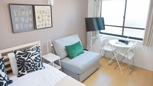 Une chambre d’hôtel avec un lit, un canapé gris agrémenté d’un coussin vert, une petite table avec une chaise, une télévision et une fenêtre avec des rideaux.