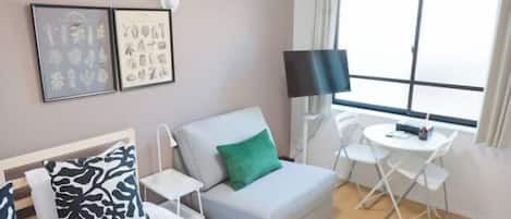 A hotel room with a bed, a grey sofa with a green pillow, a small table with a chair, a TV, and a window with curtains.