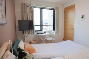 Habitación de hotel con cama, televisor montado en la pared, una mesita con sillas y una ventana con cortinas.