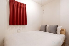 A single bed with a white mattress and grey pillows, a red curtain, and a white wall with a light switch. - Blossom Terrace Ryogoku (Tokyo)