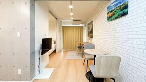 A modern living room with a TV, a dining area with a table and chairs, and a white brick wall with a painting. - Blossom Terrace Ryogoku (Tokyo)