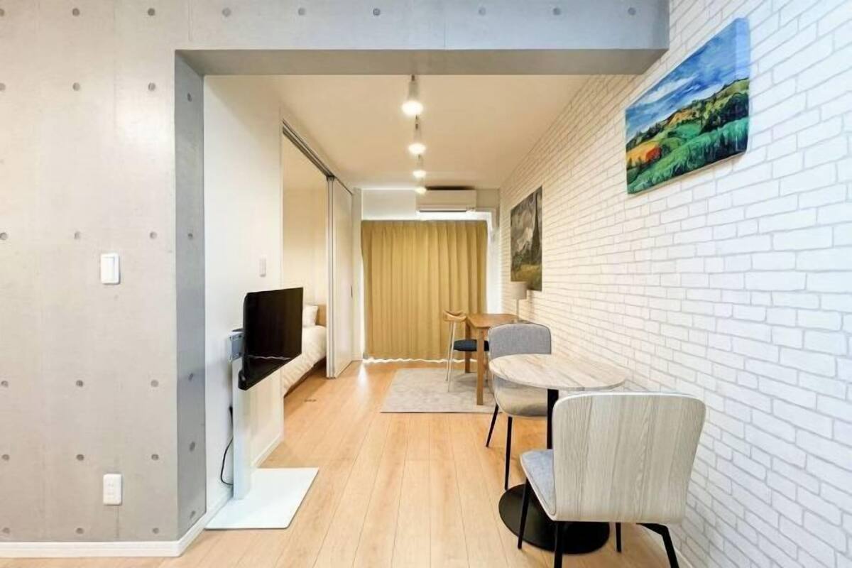 Una sala de estar moderna con televisor, un comedor con mesa y sillas, y una pared de ladrillo blanco con un cuadro.