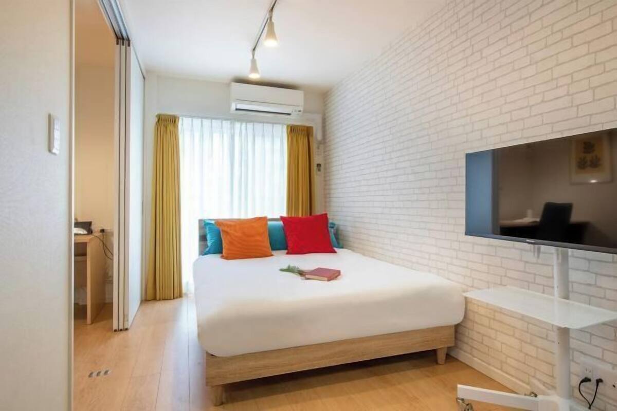 Una habitación de hotel con cama, televisión, pared de ladrillo blanco y ventana con cortinas.