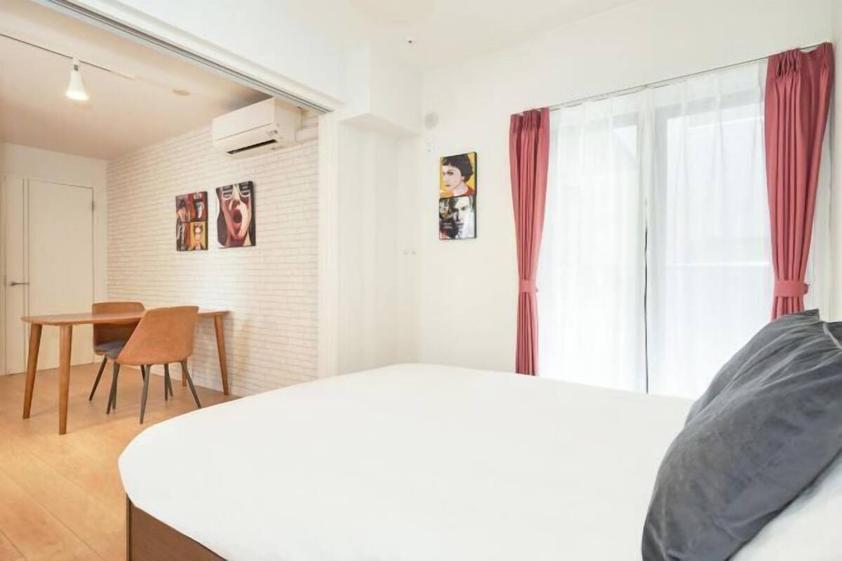 Un dormitorio con cama, escritorio y sillas, pared de ladrillo blanco y ventana con cortinas.