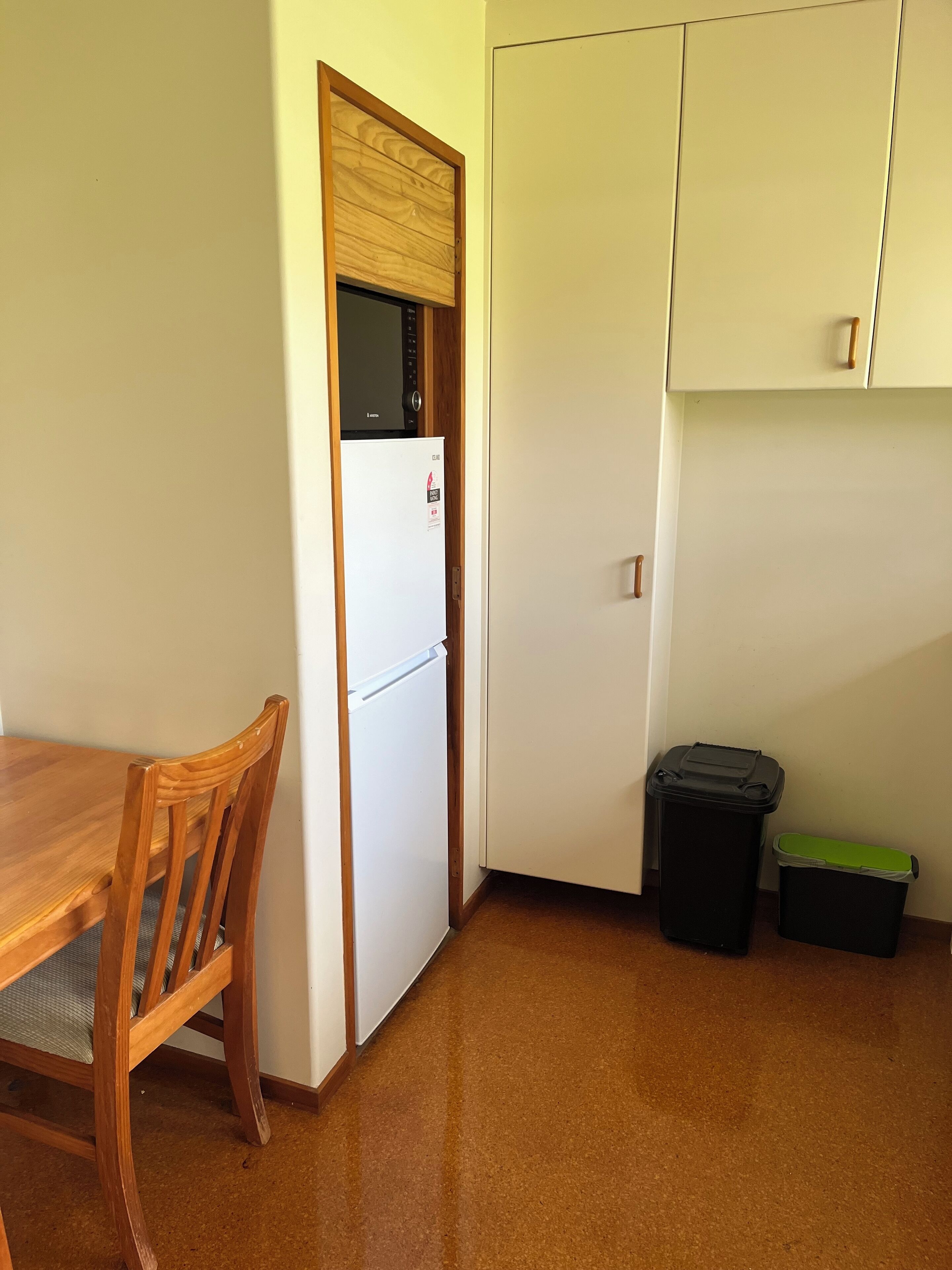 Une petite cuisine équipée d’un réfrigérateur, d’une chaise en bois, d’une poubelle et d’un placard.