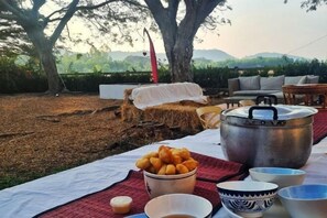 Ein Essbereich im Freien mit Tisch, Töpfen, Schüsseln und einem Korb voller Essen, vor einer Kulisse aus Bäumen und Hügeln.