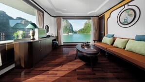 A modern room with a wooden floor, a sofa, a coffee table, a TV, and a large window offering a view of a lake and mountains. - Cycad Cruise (Bai Chay)