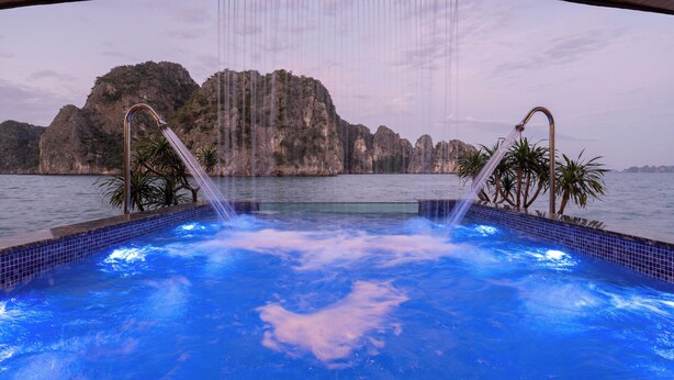 A hot tub with water jets, surrounded by a tiled pool, with a view of rocky cliffs and palm trees. - Cycad Cruise (Bai Chay)