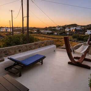 A rooftop terrace with a hammock, a blue lounge chair, and a view of a coastal village at sunset.