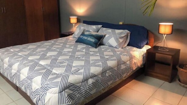 Un letto ben rifatto con una coperta a motivi geometrici, due lampade ai lati del letto e un cuscino blu con una citazione motivazionale.