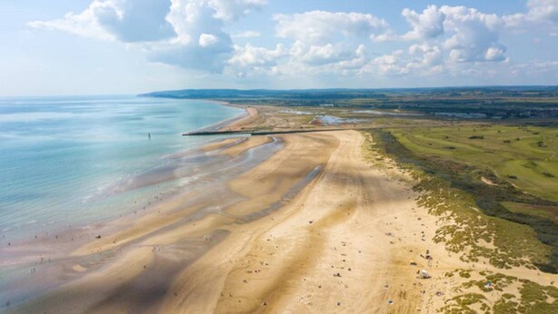 Vista aérea de una zona costera con playa arenosa, una carretera curva y campos verdes distantes.