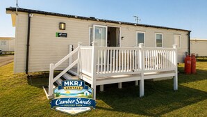 Una casa móvil con un porche blanco y una rampa, identificada con 'MKRJ Rentals' y 'Camber Sands Holiday Park'.
