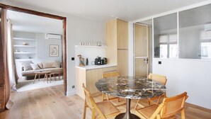 A modern living space with a round marble table, wooden chairs, a sofa, and a kitchen area with cabinets and a countertop.