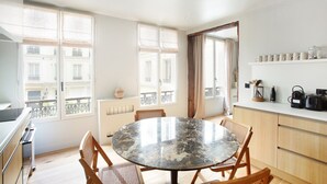A modern kitchen with a round marble table, wooden chairs, and a view of buildings through large windows.