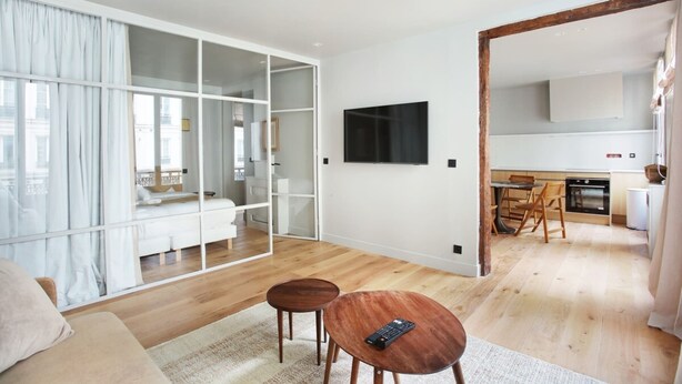 A modern living room with a sofa, two small wooden tables, a TV, and a sliding glass door leading to a bedroom.