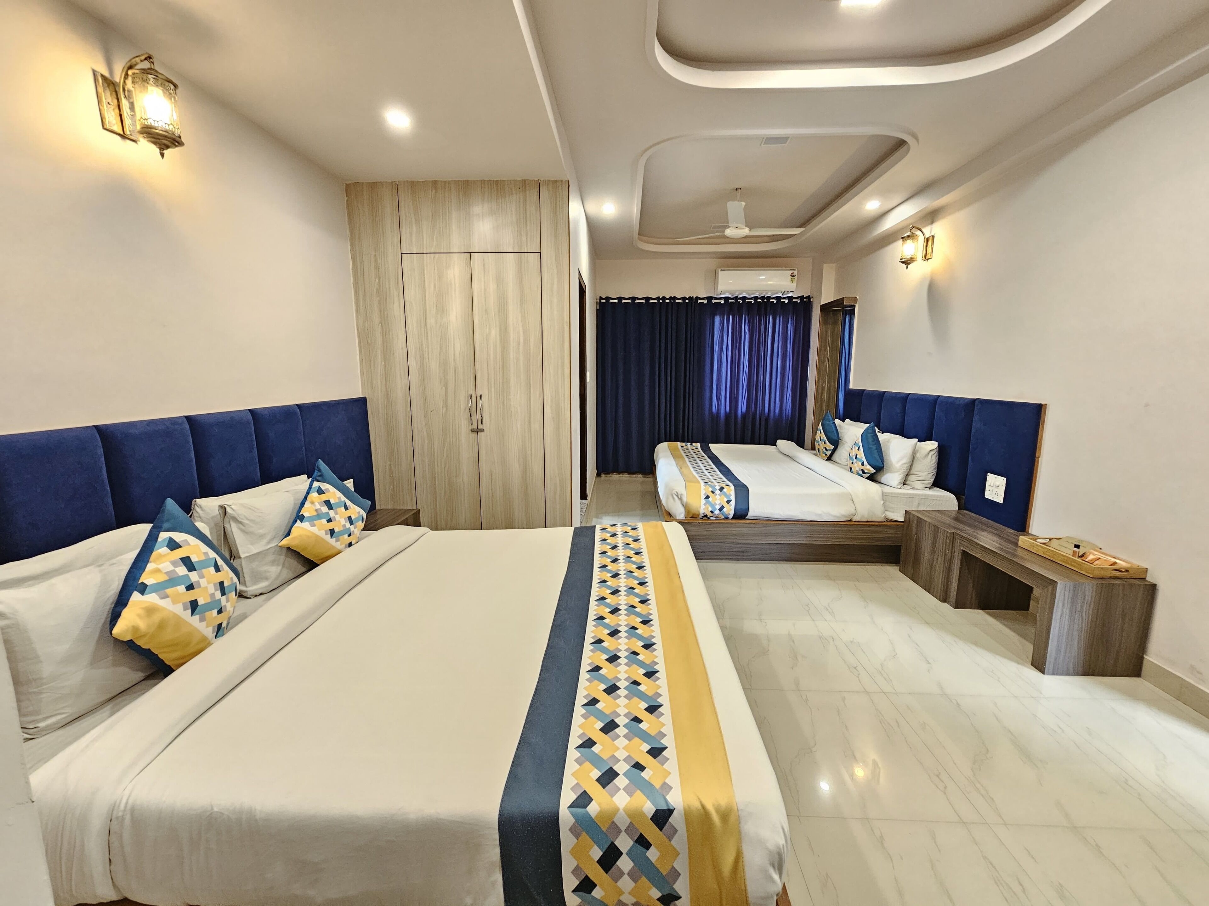 A modern hotel room with a large bed, a smaller bed, a wooden wardrobe, and a desk with a chair.