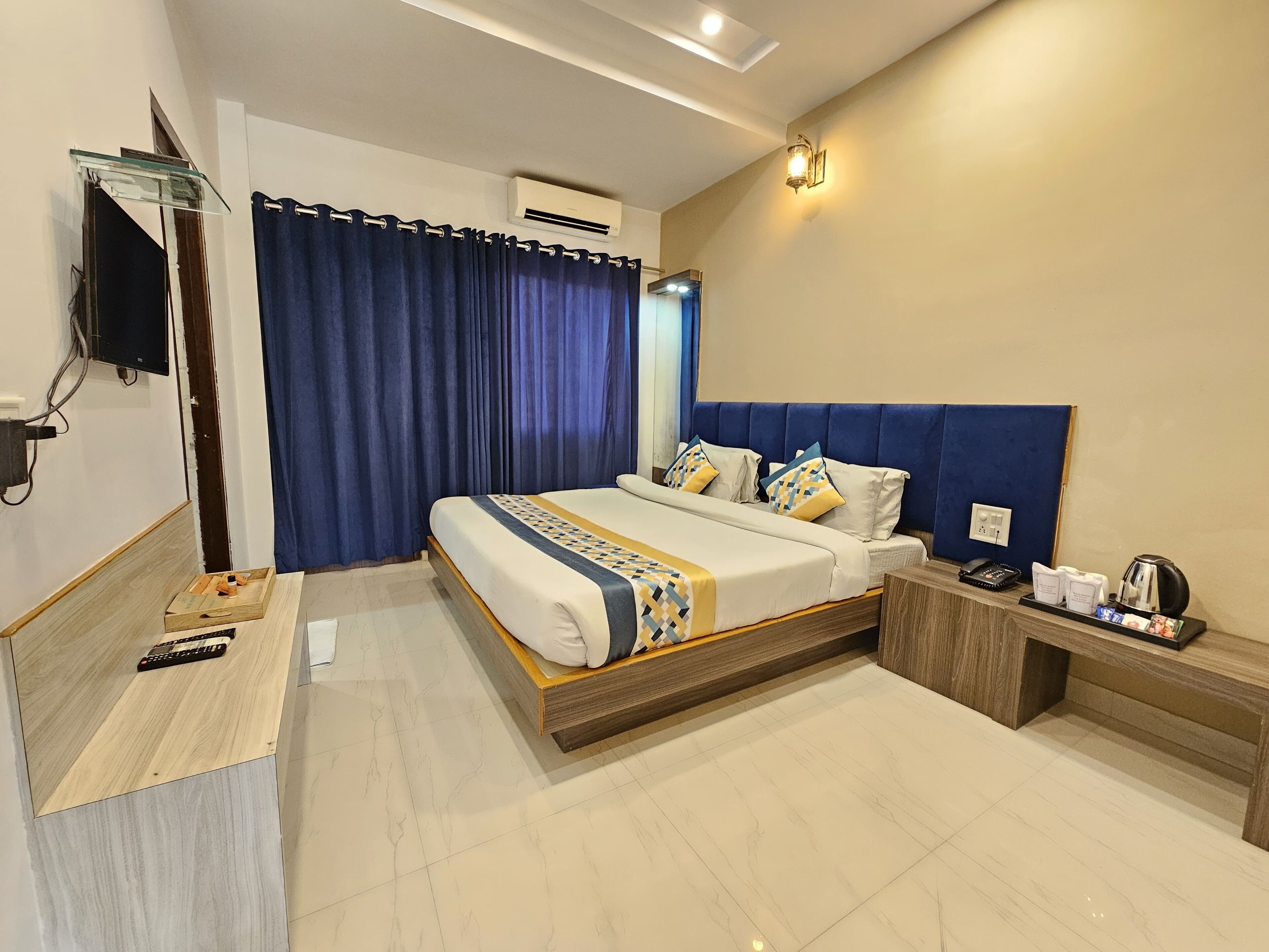 A modern hotel room with a large bed, a bedside table with a kettle, a flat-screen TV, and a large window with curtains.