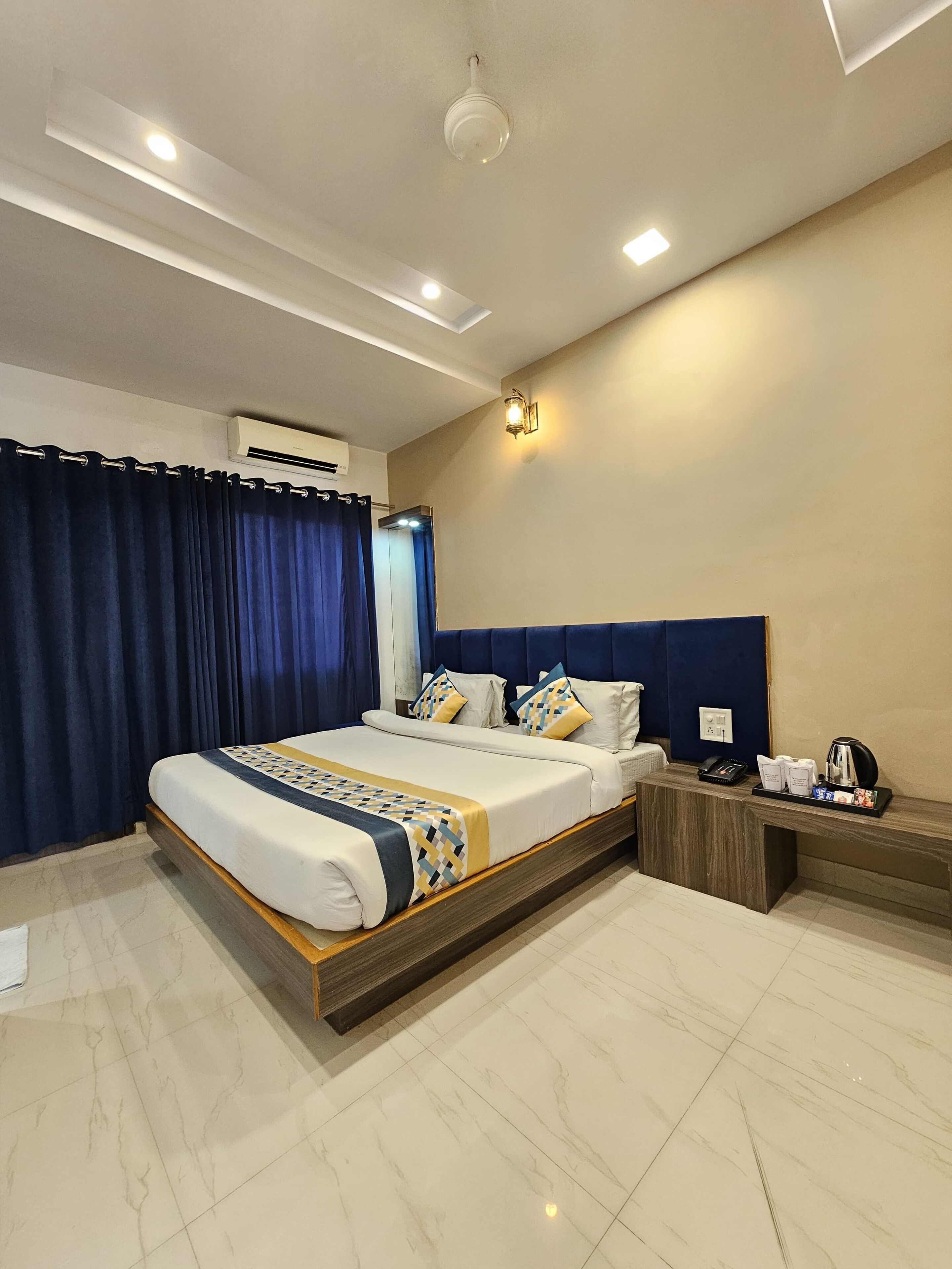 A modern hotel room with a large bed, a nightstand, a coffee maker, and a wall-mounted air conditioner.