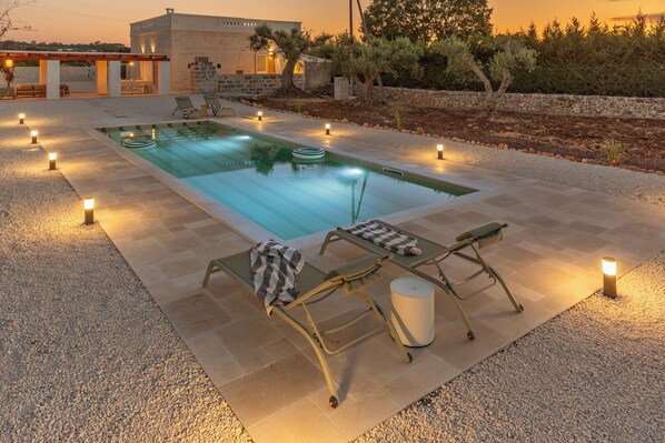 A swimming pool with lounge chairs and a small table, surrounded by a pebble path and outdoor lighting.