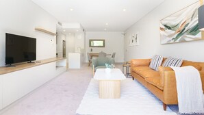 A modern living room with a flat-screen TV, a wooden coffee table, a leather sofa, and a dining area in the background.