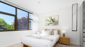 A modern bedroom with a large bed, a nightstand, a painting, and a view of trees through a window.
