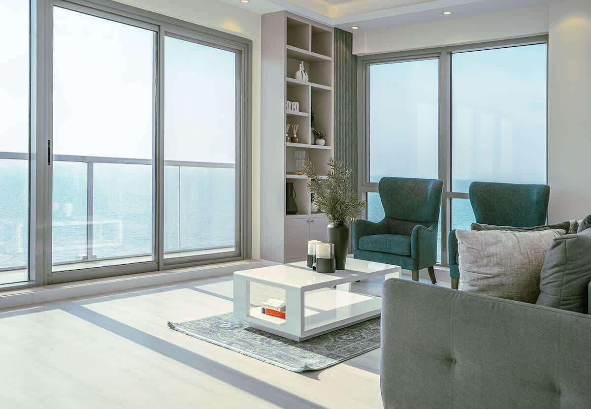 A modern living room with large windows, a white coffee table, a bookshelf, and two green armchairs.