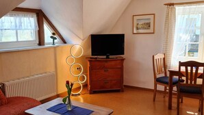 A cozy living room with a television, a wooden cabinet, a dining table with chairs, and a radiator. - Rohrdachhus- vacation apartment 2 (Mönchgut)