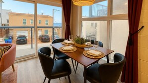 A dining area with a table set for four, a large window with curtains, and a view of the street outside. - Ocean View Balcony | 4 Min Walk to Beach | Romantic Theme (Manhattan Beach)