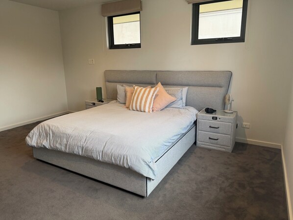 A bedroom with a bed, two bedside tables, a nightstand, a wall-mounted fan, and two windows. - Luxurious Coastal Retreat in Black Rock With Unmatched Comfort (Melbourne)