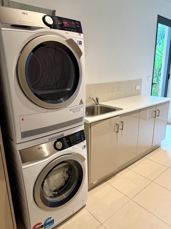 A stacked front-loading washer and dryer unit in a kitchen with a sink and cabinets. - Luxurious Coastal Retreat in Black Rock With Unmatched Comfort (Melbourne)
