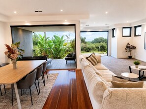 A modern living room with a wooden dining table, grey chairs, a beige sofa, and a small round table with a plant. - Luxurious Coastal Retreat in Black Rock With Unmatched Comfort (Melbourne)