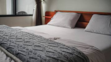 Un letto rifatto con cura, coperta e cuscini grigi, una testiera in legno e una finestra con tende.