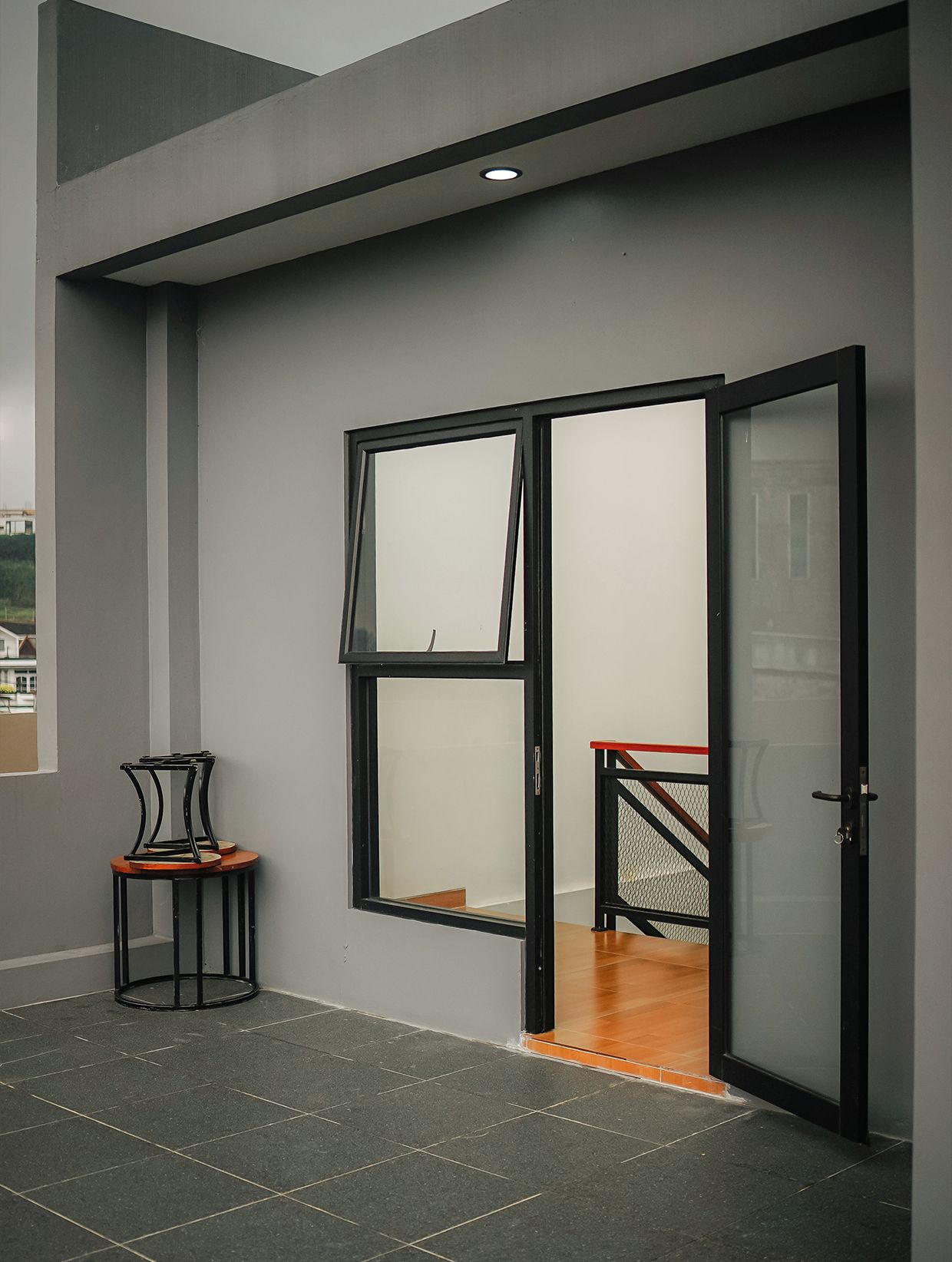 A modern hotel room with a sliding glass door leading to a balcony, a small round table with chairs, and a staircase with a metal railing.