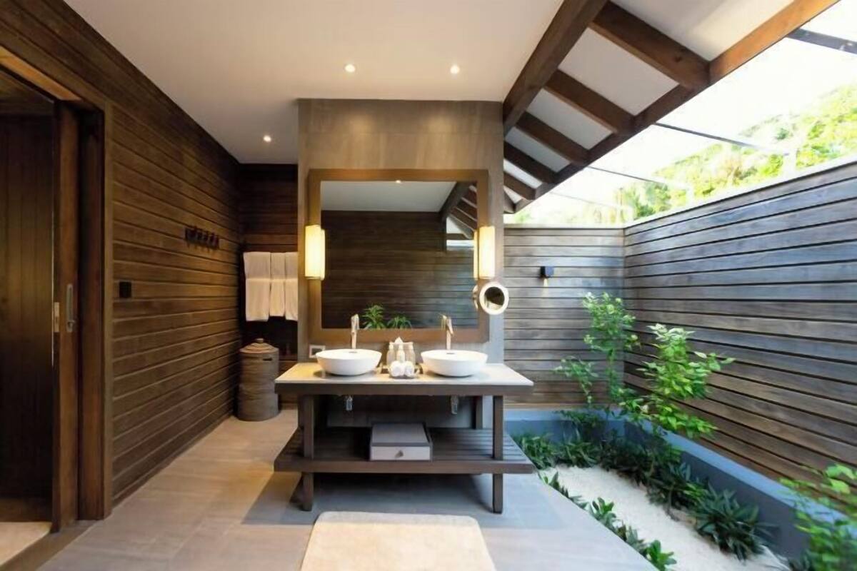 A modern bathroom with wooden walls, a central sink with two basins, a mirror, and a small plant area.