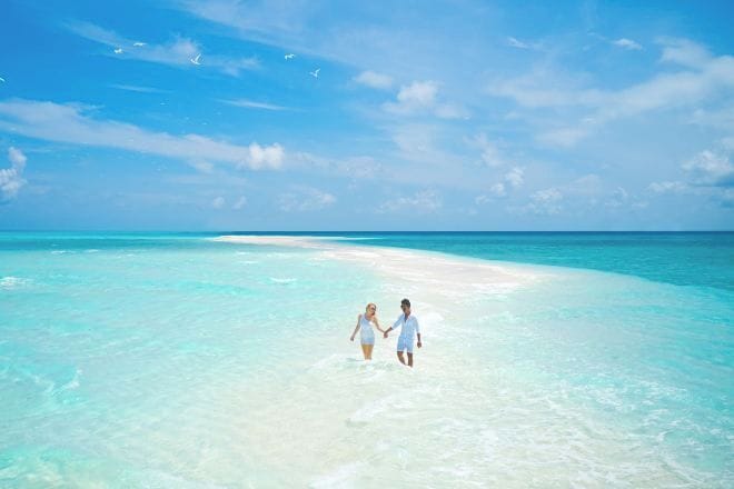 A couple walking on a beach with clear turquoise water and a sandy path.